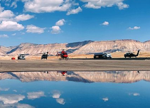 A number of helicopters are lined up on the edge of a lake reflecting the blue sky and white clouds. 