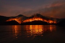 A body of water with a backdrop of silhouetted mountains with active fire burning in the distance.