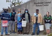 Ten volunteers holding clean up gear and in masks stand in front of the Humboldt Coastal Nature Center's concrete building. 