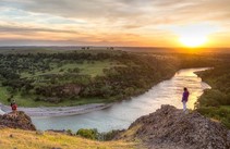 Two hikers on one outcrop with a third on another over looking a stunning sunset view of a winding river, foothills and mountains.