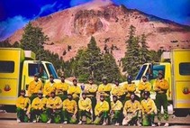 A crew of firefighters in yellow and green clothes kneeling and lined up in front of two fire engines.