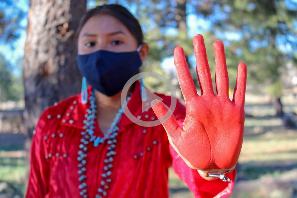 A person dressed in red with turquoise jewelry, stands and holds their red painted hand up in the stop motion 