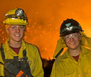 Two wildland firefighters smiling for the camera with active flames in the distance.