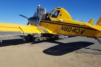 A small yellow airplane used for aerial seeding is stationed on the ground with a person standing on the wing loading seed. 