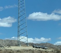 A communications tower stands tall against blue skies with two trucks in the foreground and mountains in the distance.