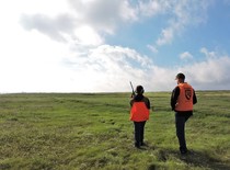 A child and man look out to into distance  of a green field, geared for hunting in bright orange vests.