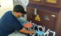  A person is bent down painting over graffiti on a trash receptacle.   