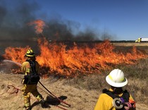 Two firefighters work on a controlled burn with active flames in tall grass.