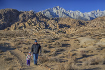 A man and a child walk along a dirt path with a snow capped mountain in the distance.