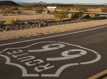 Up-close view of Route 66 with brown dirt and hills in the background, including Amboy Crater. 