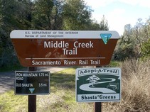 Photo of a trail sign that reads, "Middle Creek Trail, Sacramento River Rail Trail" with the Sacramento trail in front of oaks, and shrubs.