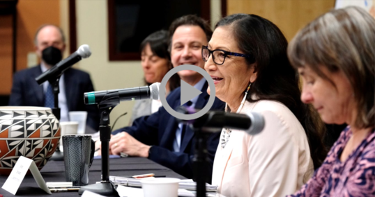 Secretary Haaland, sitting down and speaking into a microphone, smiles at a New Mexico community meeting 