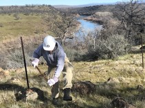 A person with a shovel digging in the ground with a river and mountains in the background.