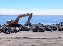 An excavator on large boulders next to the ocean.