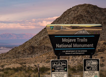 A sign that reads Mojave Trails National Monument in front of mountains at sunset.