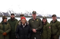 Secretary Haaland stands with eight uniformed employees in front of open water and snow capped mountains. 