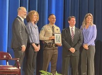 Five people standing on a stage posing for a photograph, while one is a law enforcement officer holding a plaque.