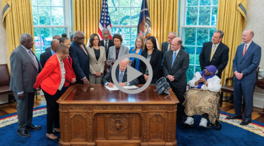President Biden signs a bill on the desk in the Oval Office as the Vice President, Secretary Haaland, and many others look on