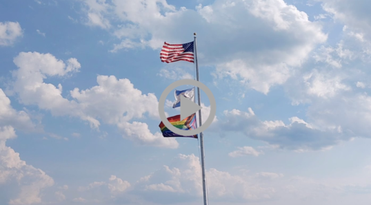 The U.S. flag, Interior flag, and Pride flag wave in the breeze against a blue sky fiiled with puffy clouds