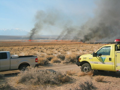A fire truck parked in a field with fires burning in the distance.