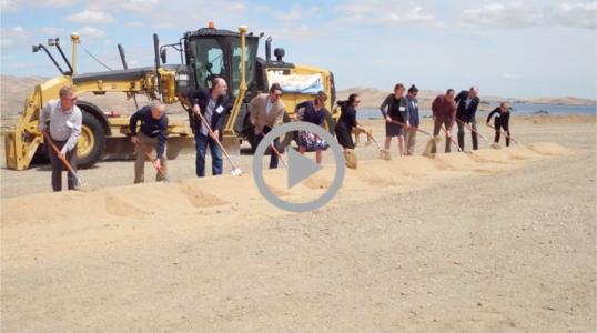 A line of people with shovels dig into the dirt at a groundbreaking ceremony with a construction vehicle behind them 