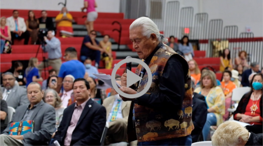 Older Native man speaks into microphone inside a crowded gymnasium