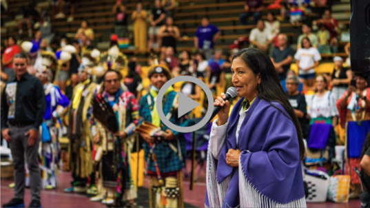 Secretary Haaland wears a blue shawl and holds a microphone in crowded gymnasium; Native Americans in traditional dress look on