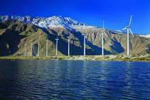 Wind turbines next to water and a mountain.