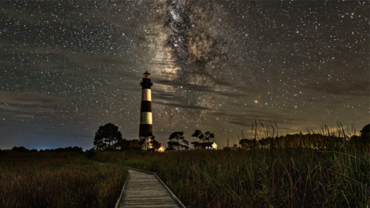 Boardwalk leading the way to a black and white lighthouse with the Milky Way rising behind it.