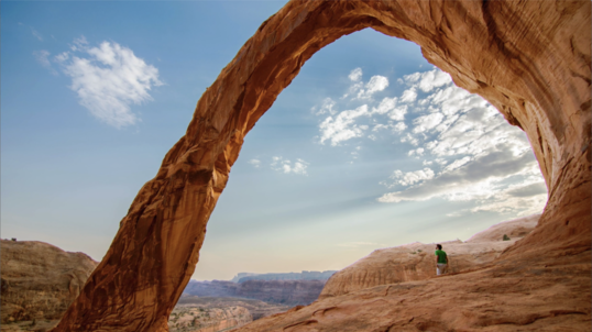 A person standing in awe under a large stone arch during the day