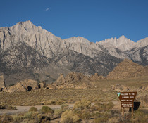 Landscape view of granite ridged mountains, a road runs through the foreground with a sign that says, "Welcome to the Alabama Hills."
