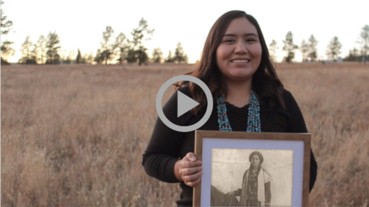 A young Indigenous woman holds up a framed black & white photograph of an ancestor