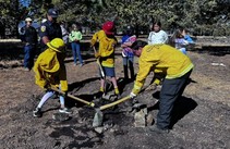 Kids in firefighter outfits use tools to put out a fire.