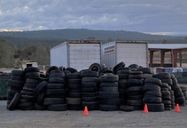 A large pile of old tires sits in front of two shipping containers and a large garage, mountains in the background. 