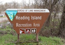 A Bureau of Land Management sign that reads, "Reading Island Recreation Area" sits on green grass with trees in the background. 