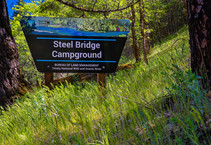 A Bureau of Land Management site sign reads, "Steel Bridge Campground" sits on a grass slope with trees surrounding it. 