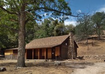 A small cabin with its windows boarded up sits in front of a hill and is surrounded by oaks. 