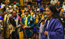 Secretary Haaland stands with a mic in her hand and a purple dance shawl on while others stand in the background in traditional regalia looking on. 