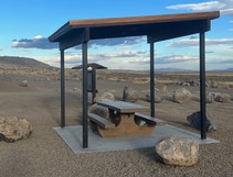 A picnic table with a shade structure above it in a gravel parking lot.