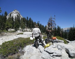Two people setting up communications in the mountains, a taller peak in the background. 