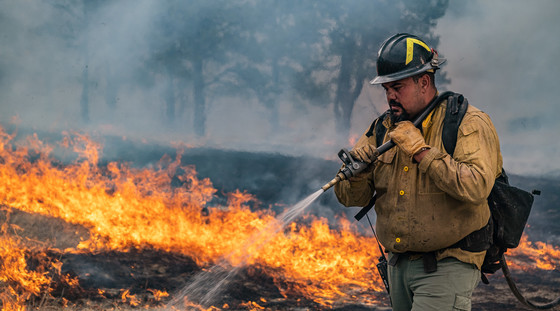 A wildland firefighter sprays water on orange-yellow flames. Smoke obscures the scorched ground and trees behind him. Photo by Colby Neal, BLM.