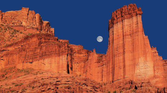 Dark red sandstone Fisher Towers and Moon in late afternoon glow. 
