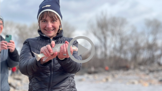Assistant Secretary for Water and Science Tanya Trujillo stands at the water’s edge, smiling and holding a fish. 