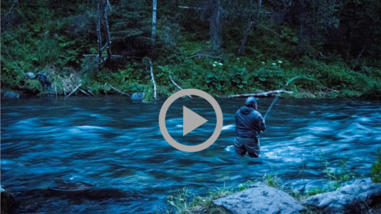 A man stands knee deep and casting his fishing rod in a flowing river banked by a steep forest.