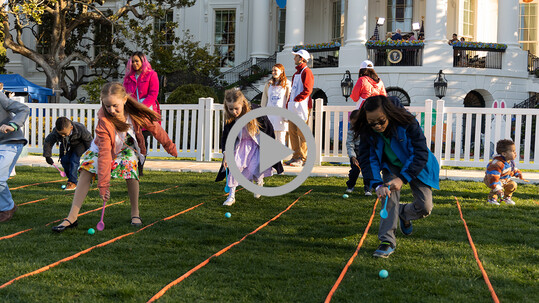 Children roll eggs on a green White House lawn with onlookers and the president giving an interview in the background