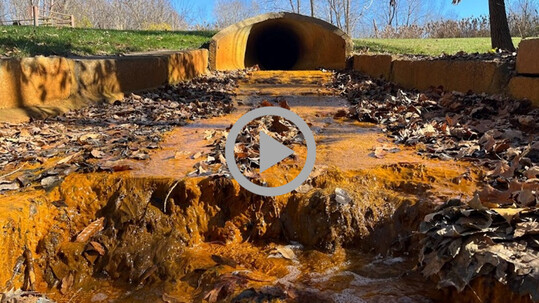 Orange, dirty runoff water trickles from a low culvert down a rocky bed