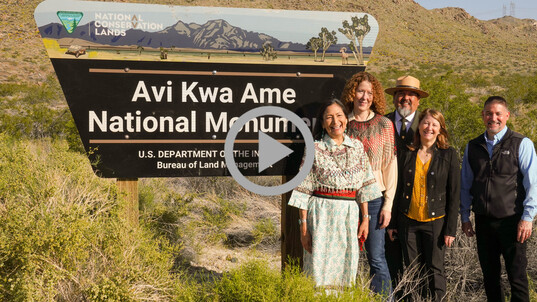 Secretary Haaland smiles and stands by the Avi Kwa Ame National Monument sign with other Department leaders.