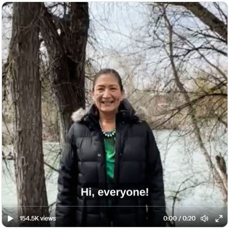 Secretary Haaland smiles and stands in front of several trees by a river