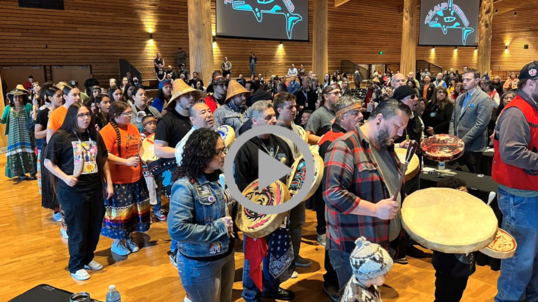 A procession of Native Americans singing and playing drums in a large auditorium. 