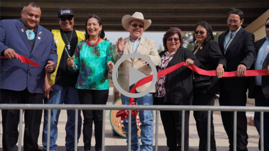 Secretary Haaland cuts the ribbon on a new bridge for the Mescalero Apache Tribal community, as smiling members of the Tribe look on and applaud. 